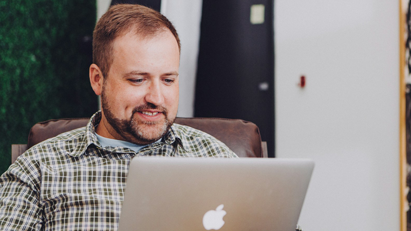 A man smiles as he sits in a chair and uses his MacBook.