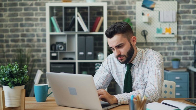 Frustrated employee working on a laptop at this desk after being required to attend an unpaid pre-shift meeting.