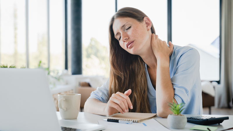 woman in blue shirt rubbing her neck with her eyes closed at her office desk, looking exhausted