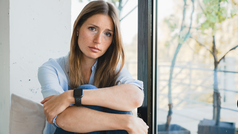 Woman sitting while hugging one knee with a drained, subdued expression