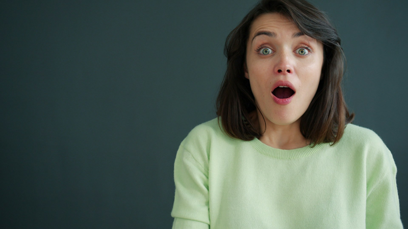 A woman has a surprised expression while standing in front of a dark background.