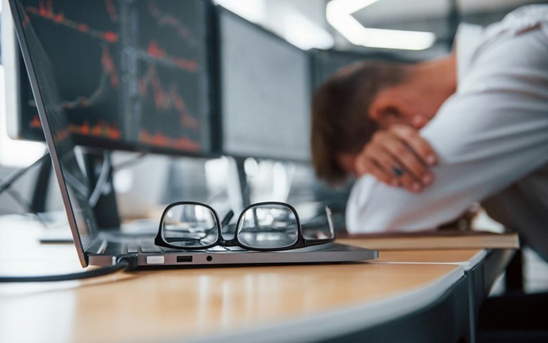 A man puts his head in his hands at his desk, his laptop in the foreground