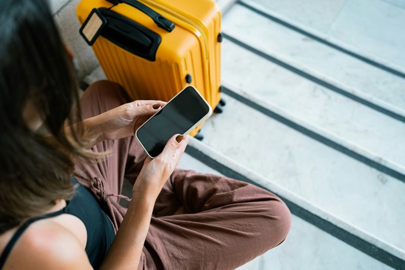 A woman looks down at her phone sitting on the ground with a suitcase next to her