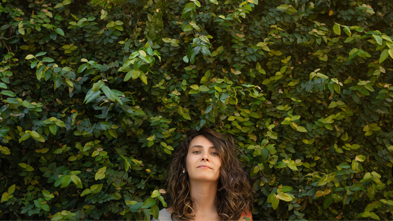 Woman stands beneath a tall, dense green hedge outdoors, looking calmly toward the camera.