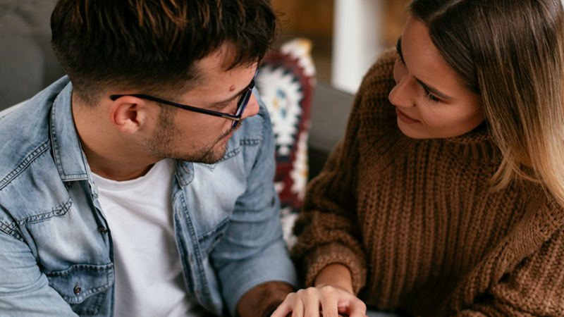 A man and a woman sitting beside one another clasp hands and look at one another.