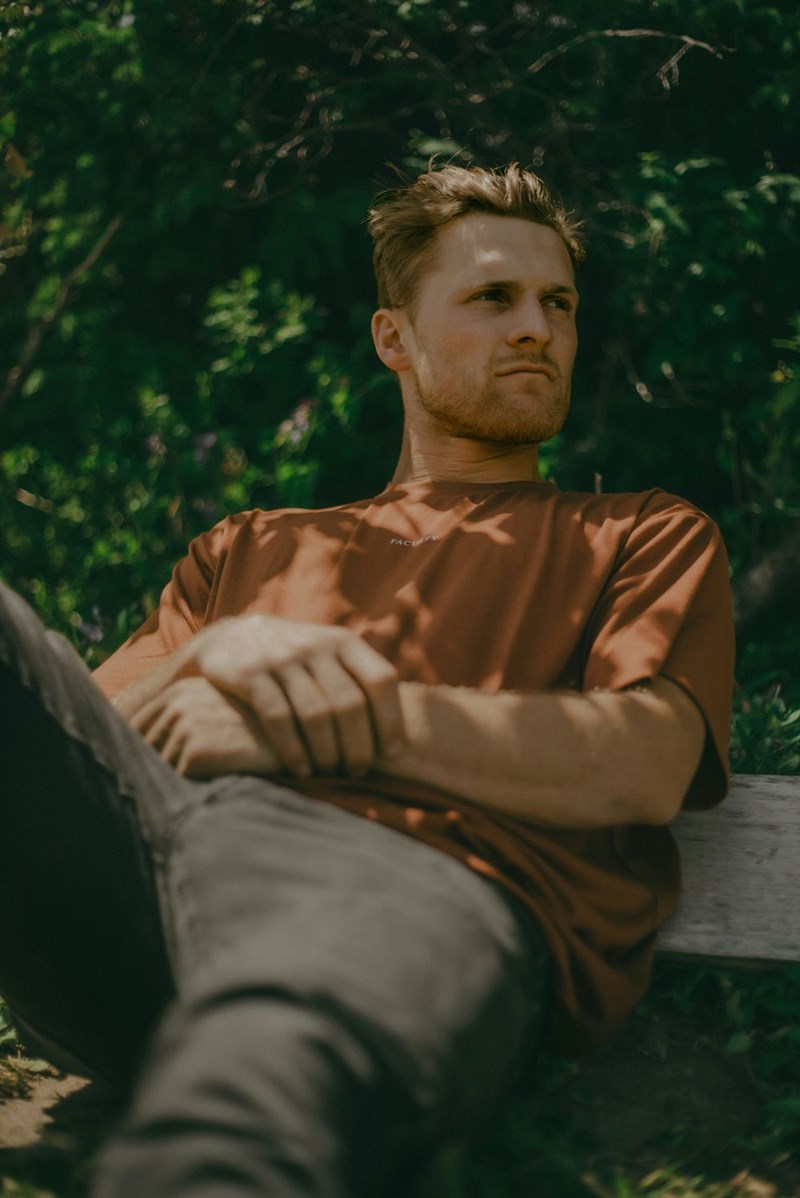 guy in a rusty orange colored t shirt sitting outdoors with his arms folded and a derious determined expression