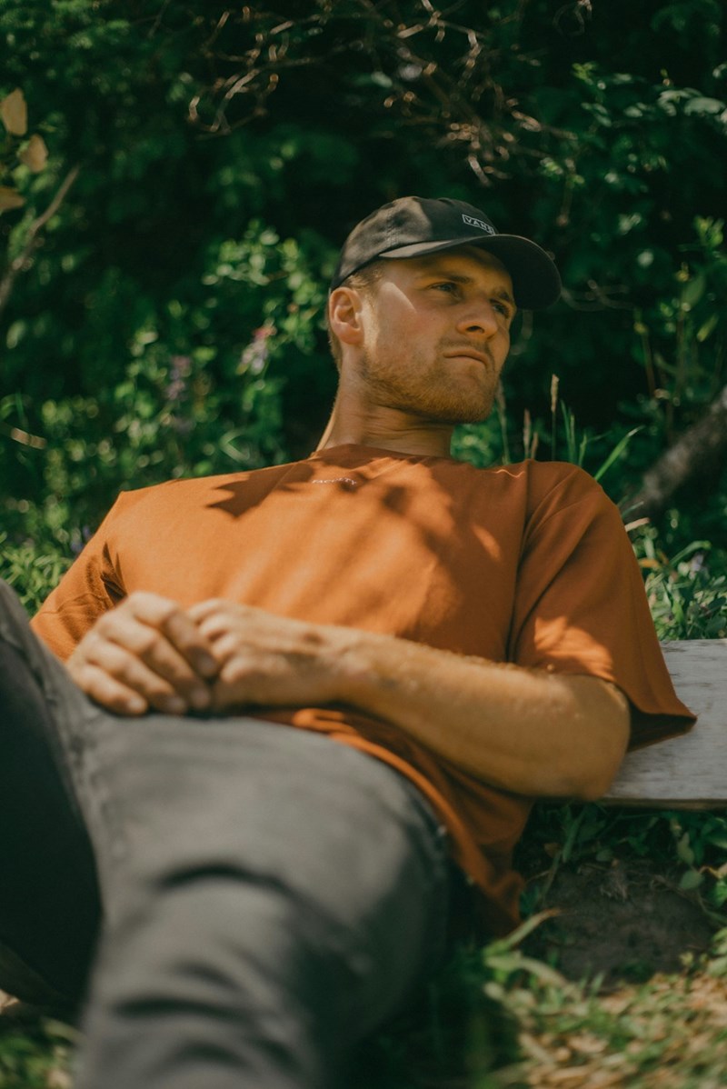 Man in a black cap and orange tshirt sitting outdoors with a concerned expression