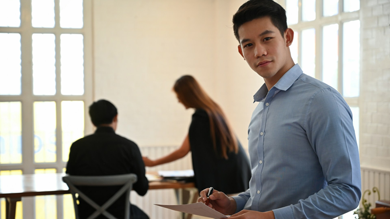 An employee stands while holding a piece of paper, with colleagues discussing something in the background.