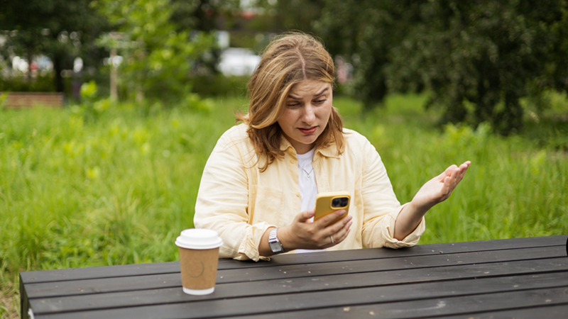 Upset woman react with confusion to something on her phone while sitting at a table in a green garden