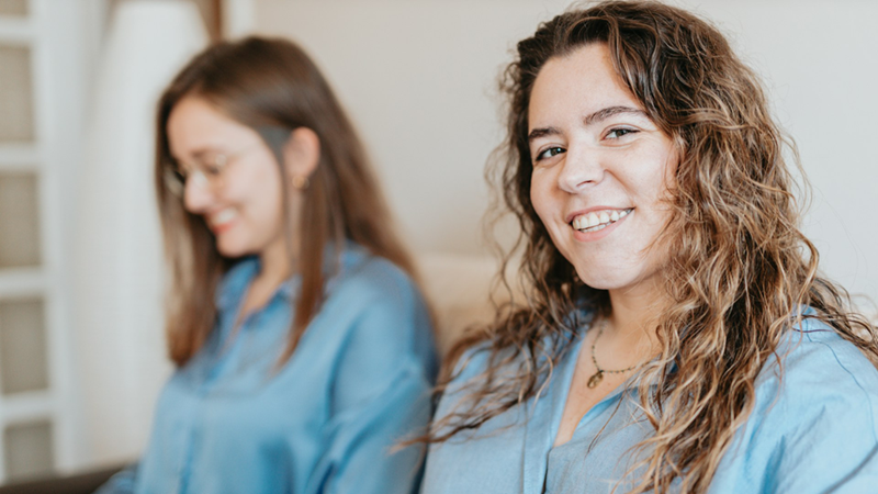 A woman smiles while sitting on the couch, her coworker blurred in the background, looking away and smiling.