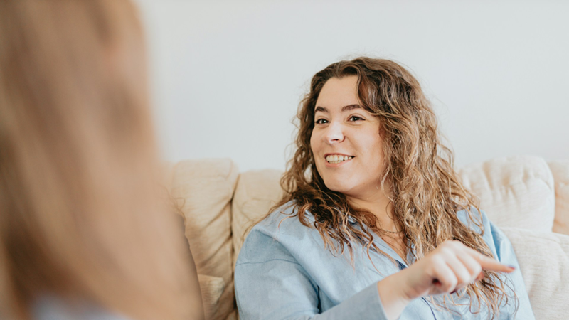 A woman sitting on a couch speaks to a colleague, who is shown by the back of her head.