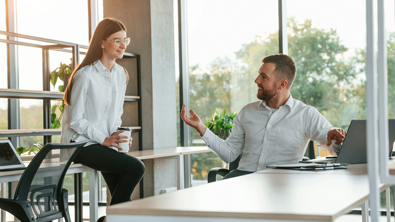 An employee sits at a desk holding a coffee cup as her boss sits opposite her, explaining something while working on his laptop.