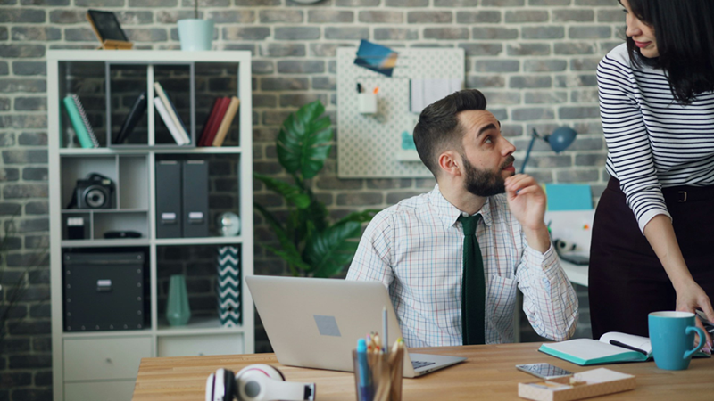 A woman leans slightly over the desk of a male colleague, looking up at her from working on his laptop.