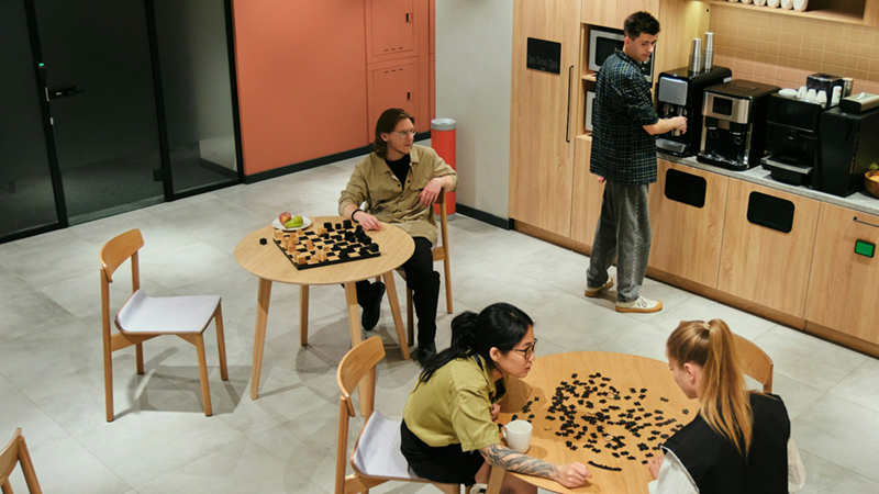 Employees socialize and prepare coffee in a break room with board games on the tables.