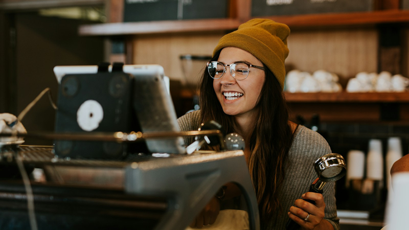 A barista wearing a beanie and glasses laughs while holding the portafilter for a coffee machine.