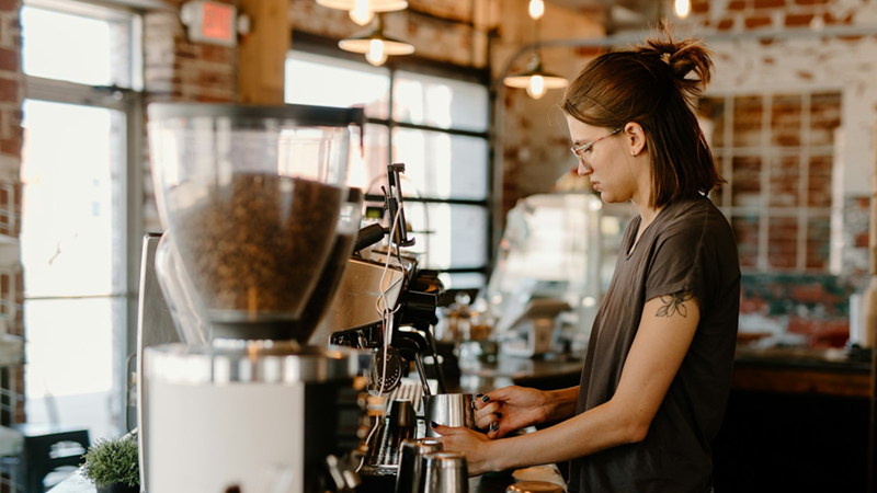 A barista with half her hair tied back heats up milk in a jug using a coffee machine.