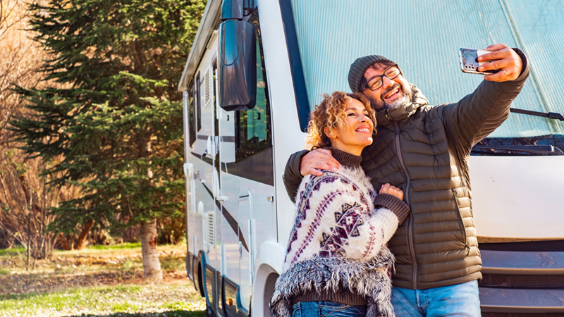 Couple smile for a selfie beside a large white camper parked among trees.