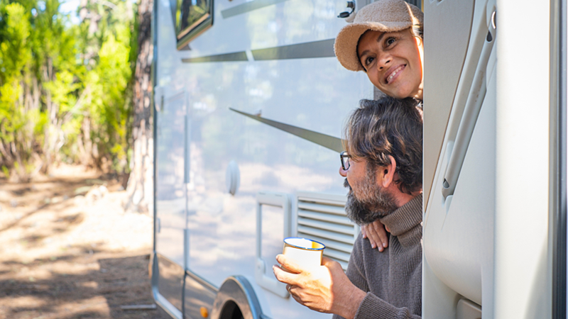Couple smile from the doorway of a white camper, one holding a mug beside a wooded area.