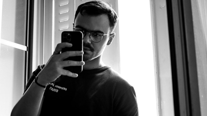 Black and white photo of a man wearing glasses, reading something on his phone with a discontented look on his face, like his reading something he doesn't understand or agree with
