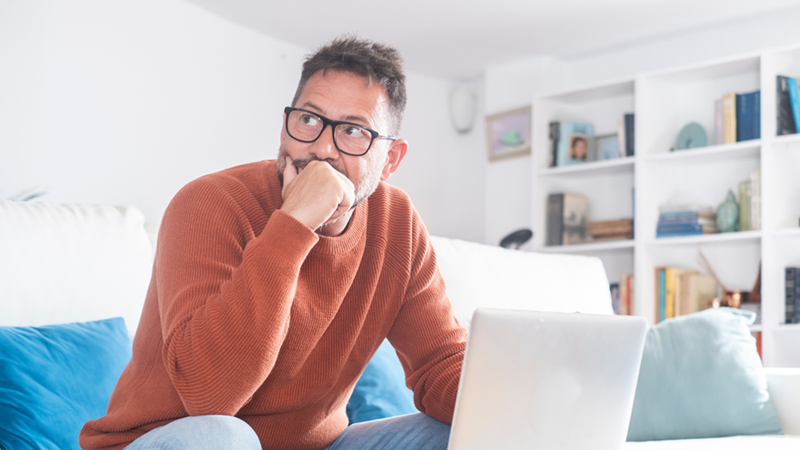 Man wearing glasses resting his hand near his mouth while sitting beside a laptop with a concerned expression. like he is troubled by something he discovered