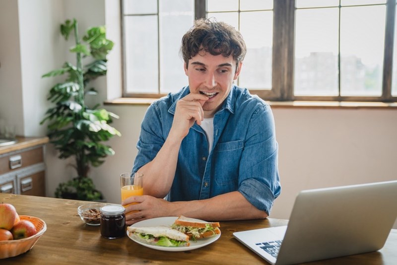 Man eating a sandwich while working from his home kitchen looking amused by something on his laptop