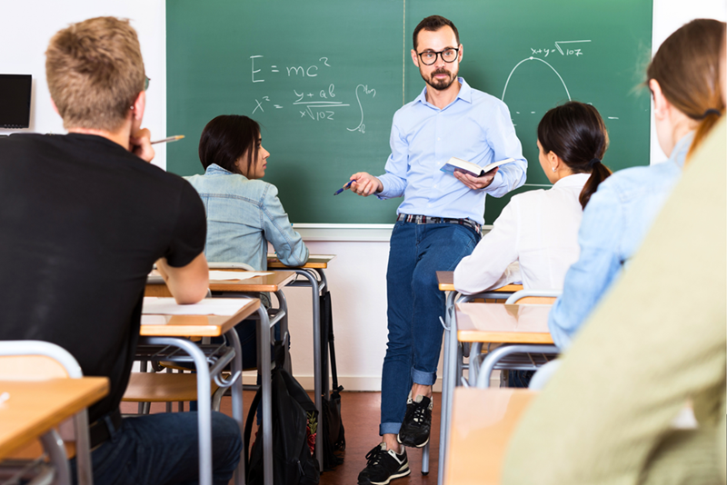 A teacher in front of a high school class, asking the students a question in a more casual manner, leaning on a table.