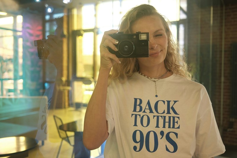 A woman takes a picture with a camera with a t shirt that says "Back to the 90s"