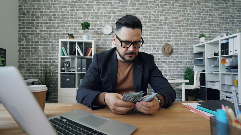 A man sits at a work desk, counting out bank notes.