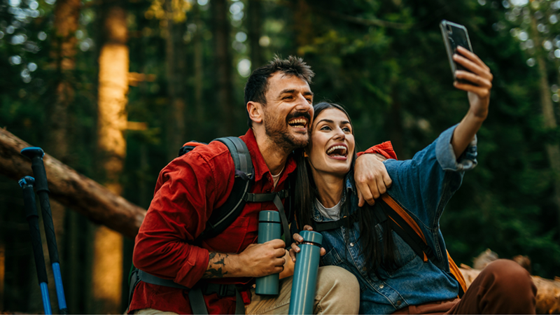 Two hikers with backpacks smile for a selfie in the woods while holding metal water bottles.