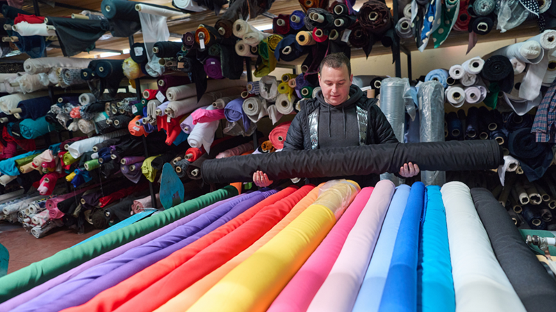 Man holds a large black fabric roll in a textile warehouse surrounded by colorful bolts of fabric as he examines a rack of fabric rolls.