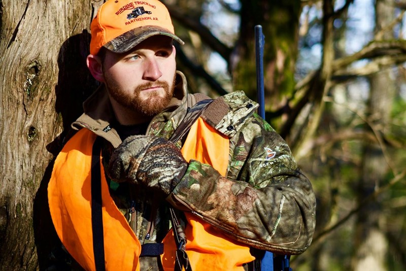 Man in hunting gear outside leaning against a tree.