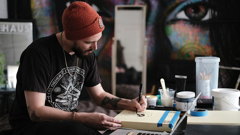 Bearded man in a beanie draws on paper at an art table surrounded by brushes, paint, and supplies.