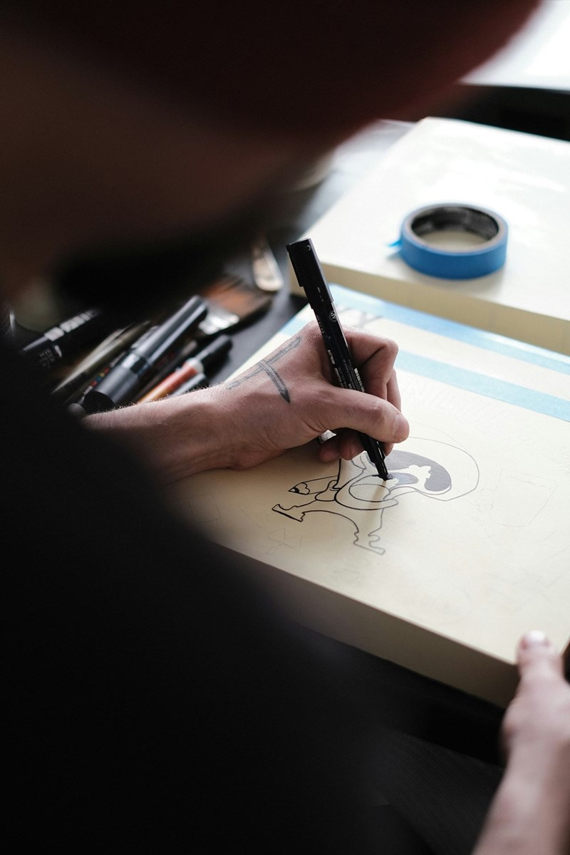 Close-up of a hand using a black marker to ink an illustration on paper beside pens and blue tape.
