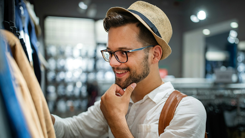 A man wearing a trilby strokes his beard while browsing in a clothing store.