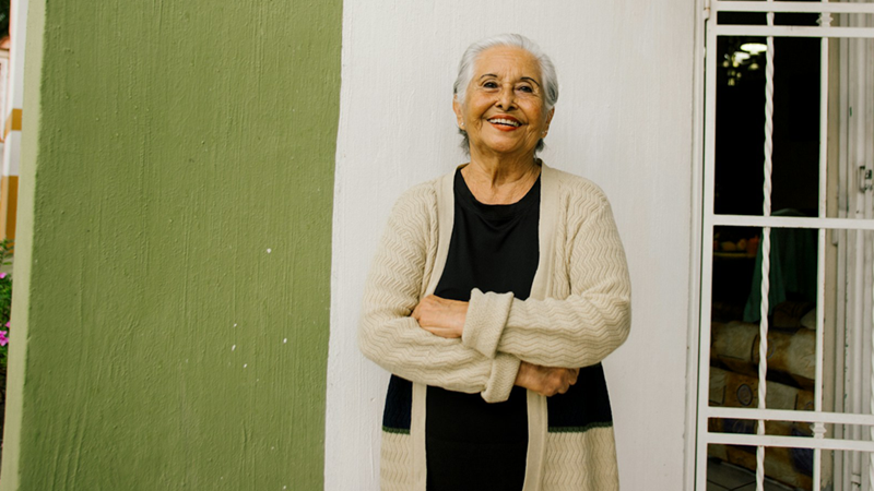An older woman smiles and crosses her arms while standing in front of her house.