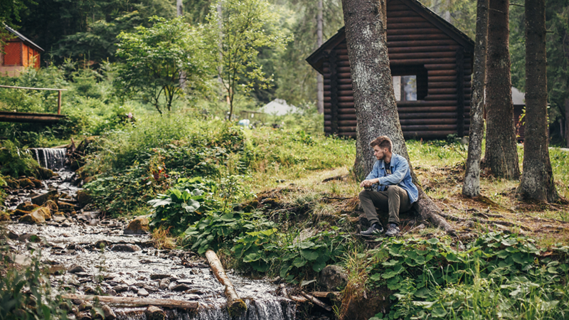 Man sits beside a forest stream near a wooden cabin, surrounded by trees and greenery.