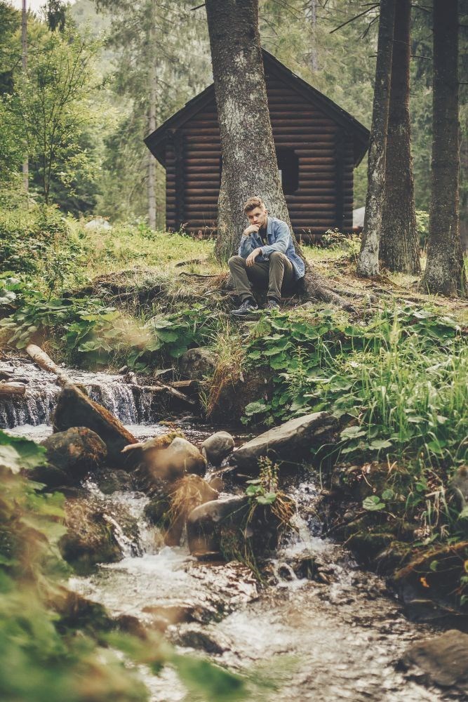 Man sits beside a forest stream near a wooden cabin, resting his chin on one hand.