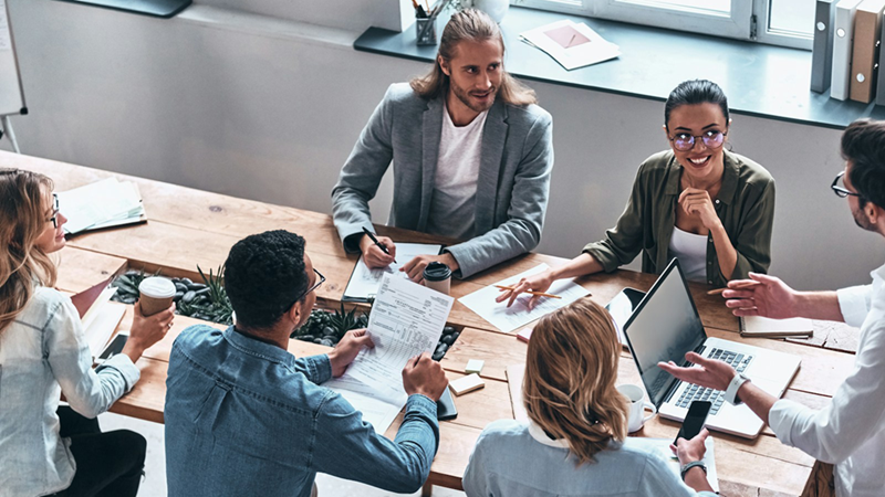 Overhead view of employees sitting around a table as a manager presents to them.