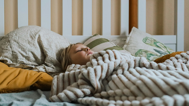 A young child sleeping on their back under a blanket in a bed with a white wooden frame.
