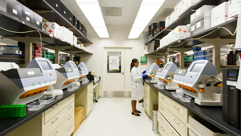 A woman in a white coat stands at the back of a lab, working on a machine.