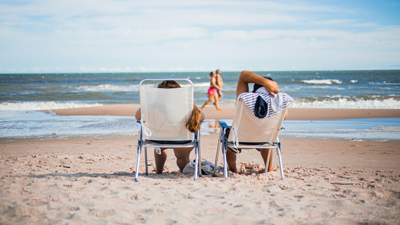 Two people relaxing in folding chairs on the beach, looking at the sea.