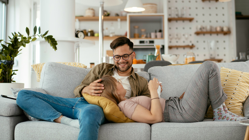 A couple relaxes on a couch, the woman resting in the man's lap with her head on a cushion.