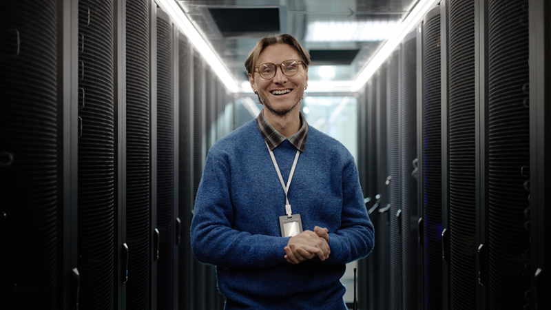 Smiling man with glasses and an ID badge stands between rows of server racks