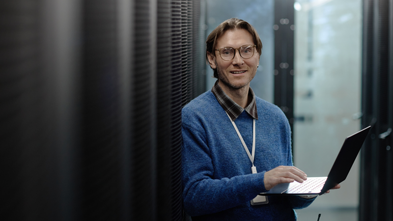 Man with glasses holds an open laptop between server racks