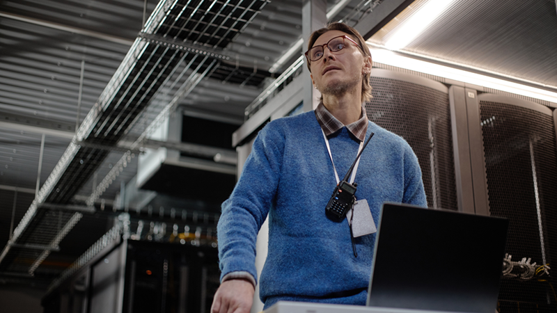 Man with glasses stands in a server room beside an open laptop with a worried expression
