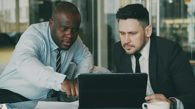 Two male coworkers sit looking at a laptop together, with one pointing at the screen.