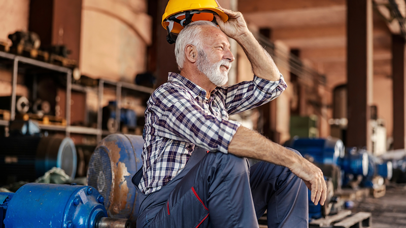 Older bearded worker smiles while lifting a yellow hard hat as he sits among industrial machinery.