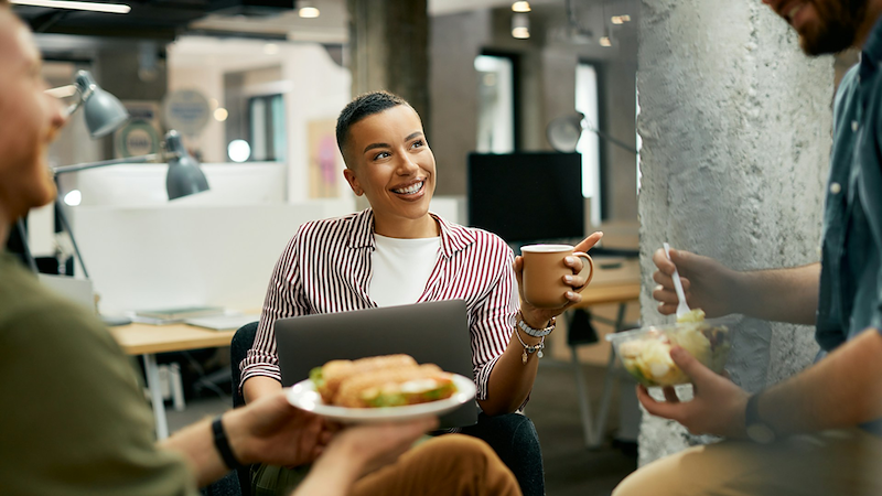 An employee holding a mug with her laptop on her lap gestures to her coworkers, who are eating lunch.