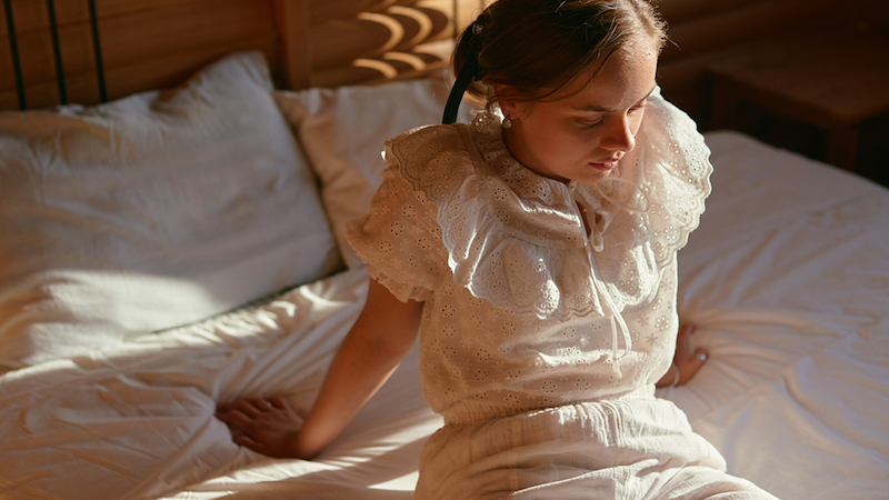 A young woman puts her hands behind her as she sits on a bed, wearing white cotton pajamas.