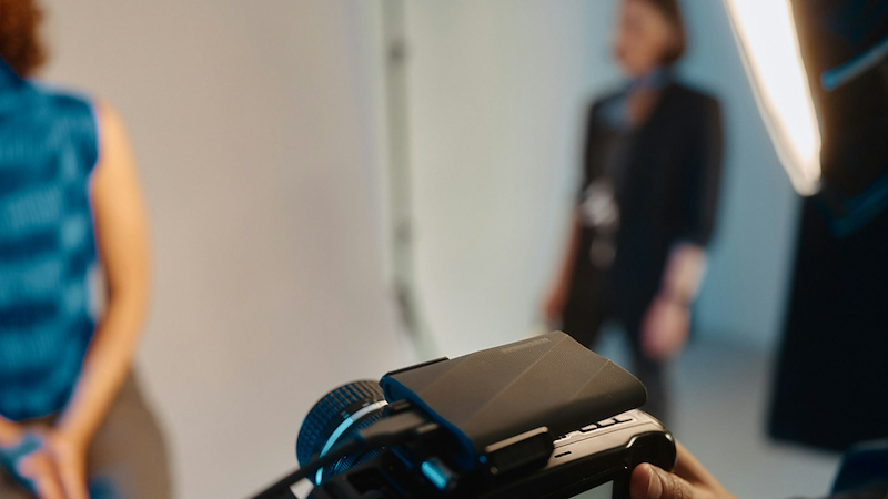A photographer points a camera at a woman, with another woman wearing a lanyard standing in the sidelines.